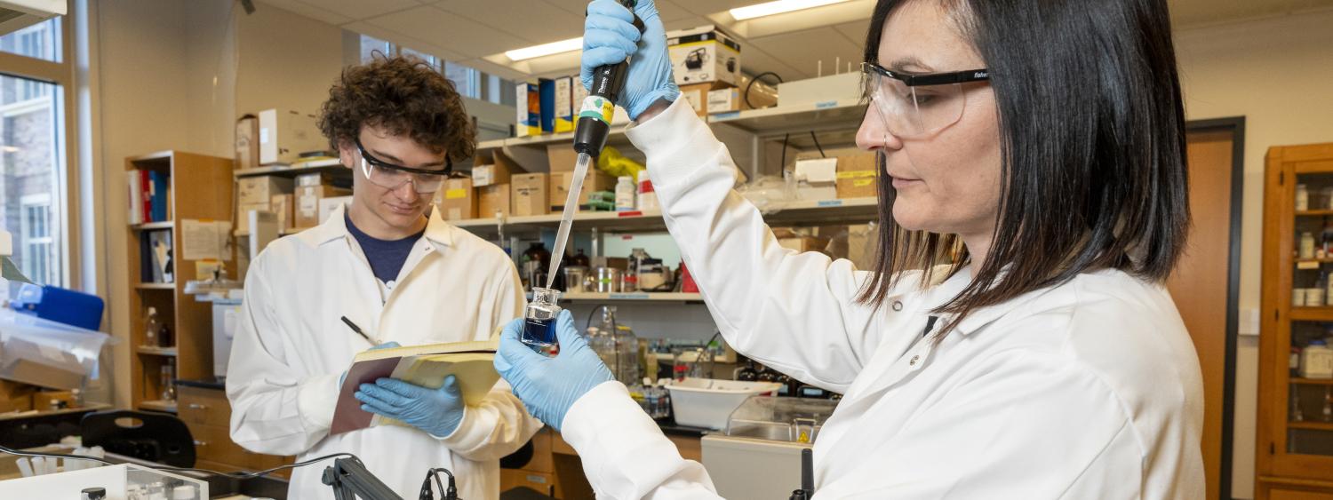 Researchers in white coats with researcher in foreground pipeting a colored liquid.