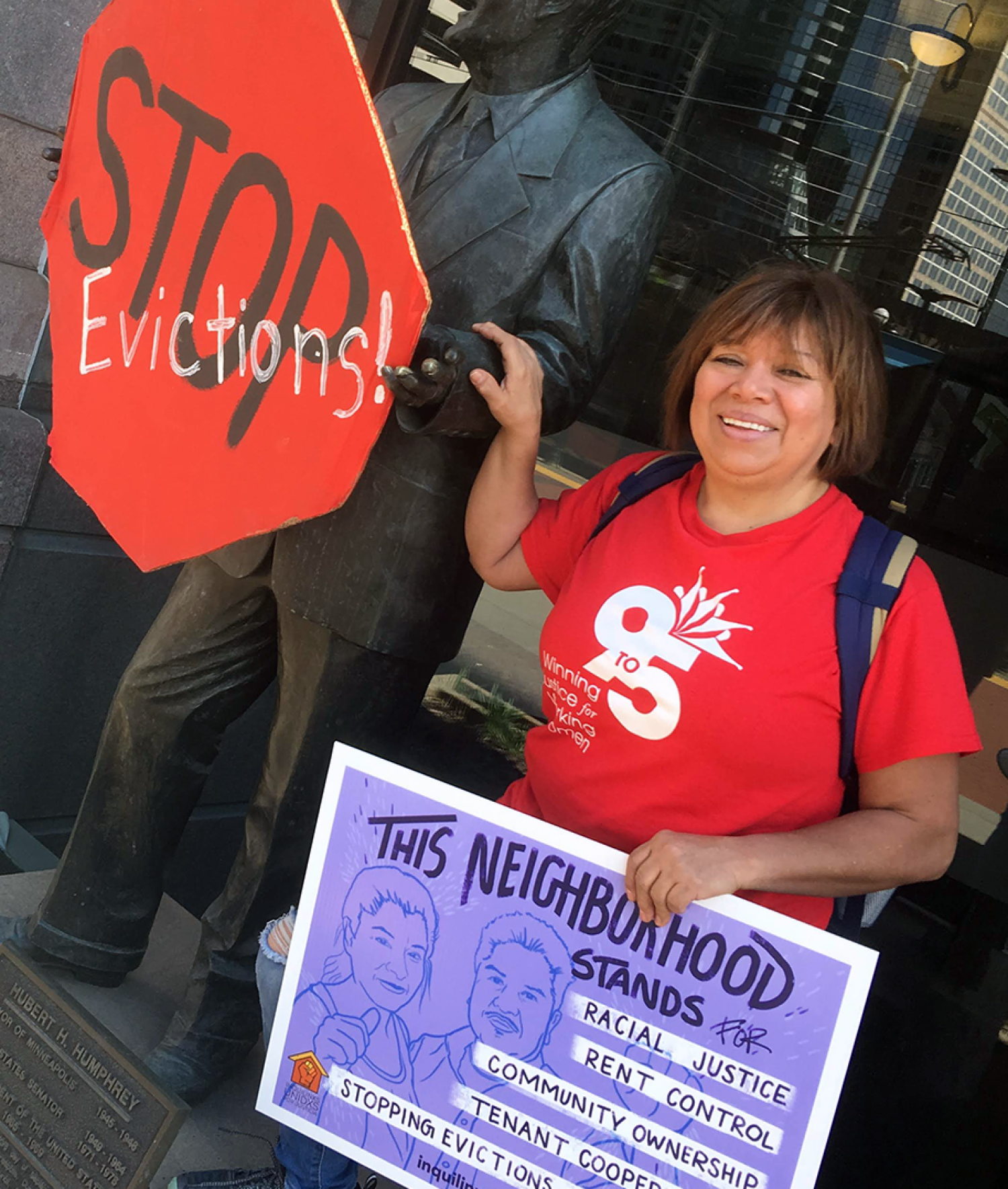 Luz Galicia stands by a statue holding a sign that says "Stop evictions." Luz is holding a sign that says this neighborhood stands for...