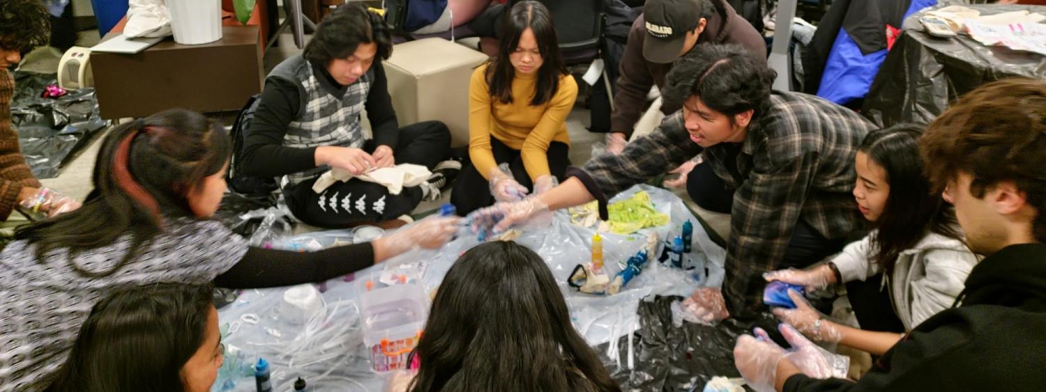 Students sitting in circle on floor tie dying tote bags with group of students around table knitting in background