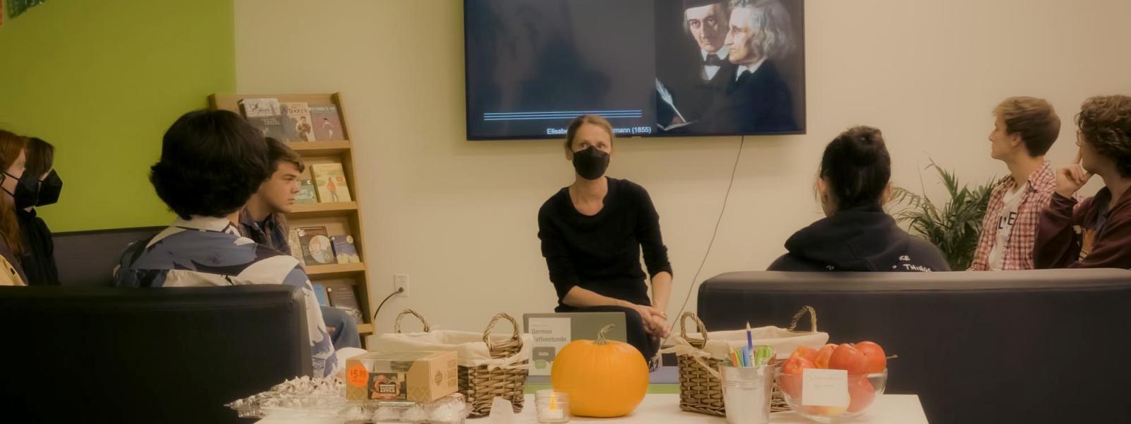 Students gathered around instructor and screen displaying Brother's Grimm slide, table with pumpkins and assorted snacks in foreground