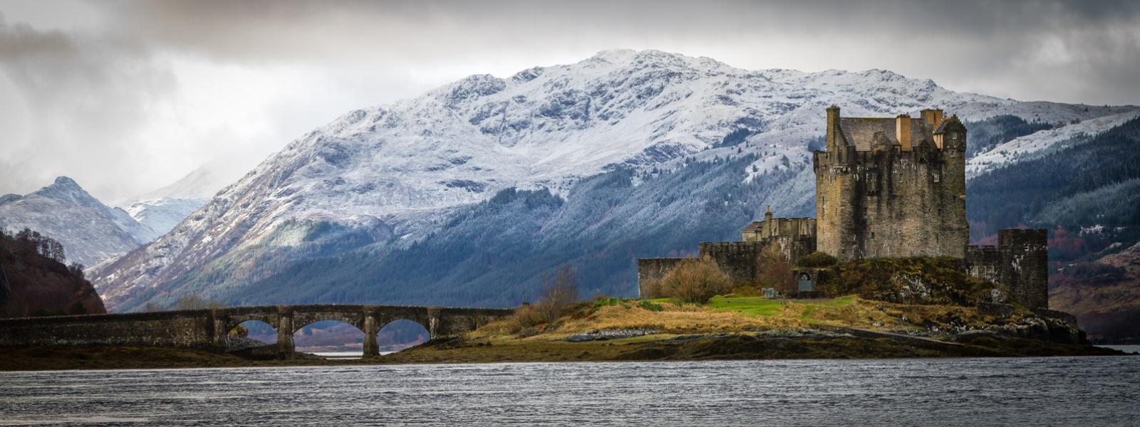 Castle with mountains in the background and a bridge leading to it.