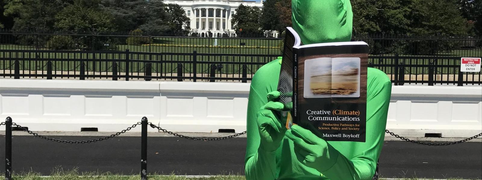 Person in green suit sitting in front of the White House reading a book, Creative (Climate) Communications by Maxwell Boykoff