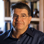 Older man with glasses and a blue shirt leaning against a wall