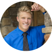 Man with blond hair learning against a pillar in a blue button down shirt and black tie