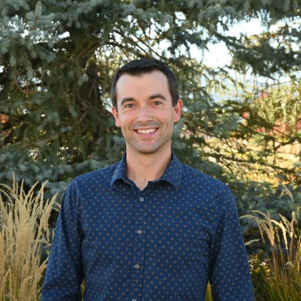 Man with brown hair smiles in front of trees
