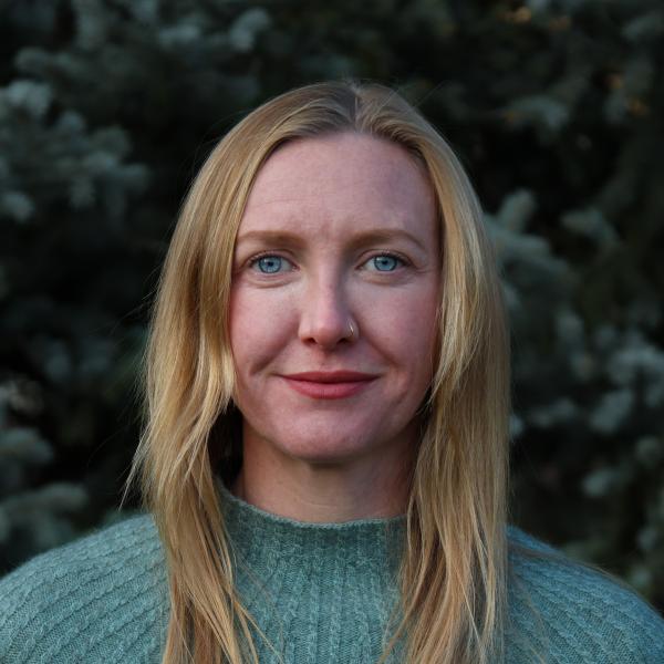 Woman with blonde hair and blue sweater stands in front of tree