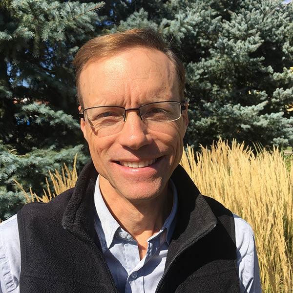 Man with glasses smiling at camera in a blue shirt and vest with pine trees in the background