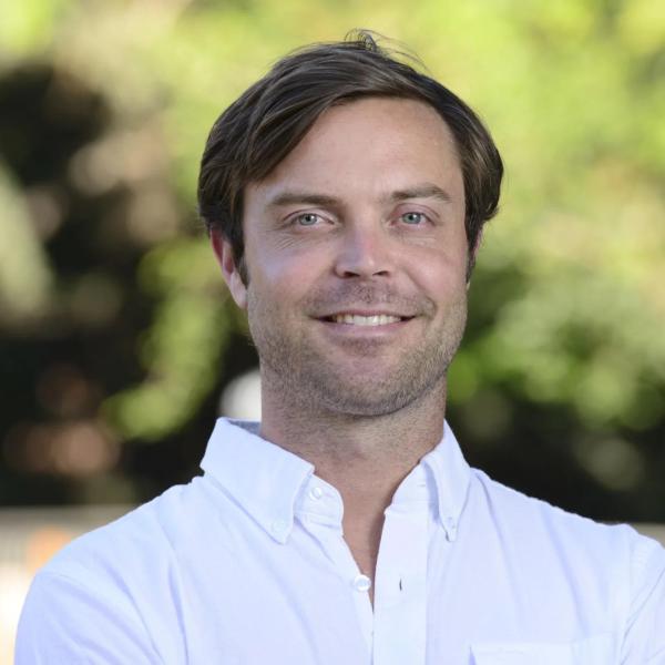 Headshot of a man with short, parted brown hair in a white collared shirt in front of a green blurred background