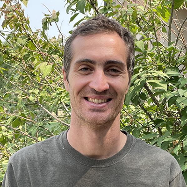 Professor in gray t-shirt standing in front of green bush