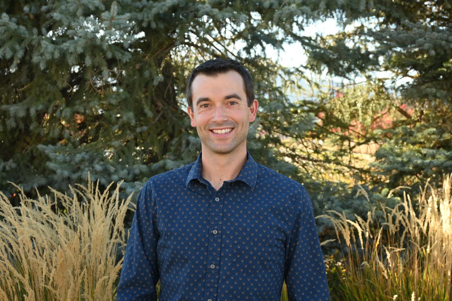 Man with brown hair smiles in front of trees