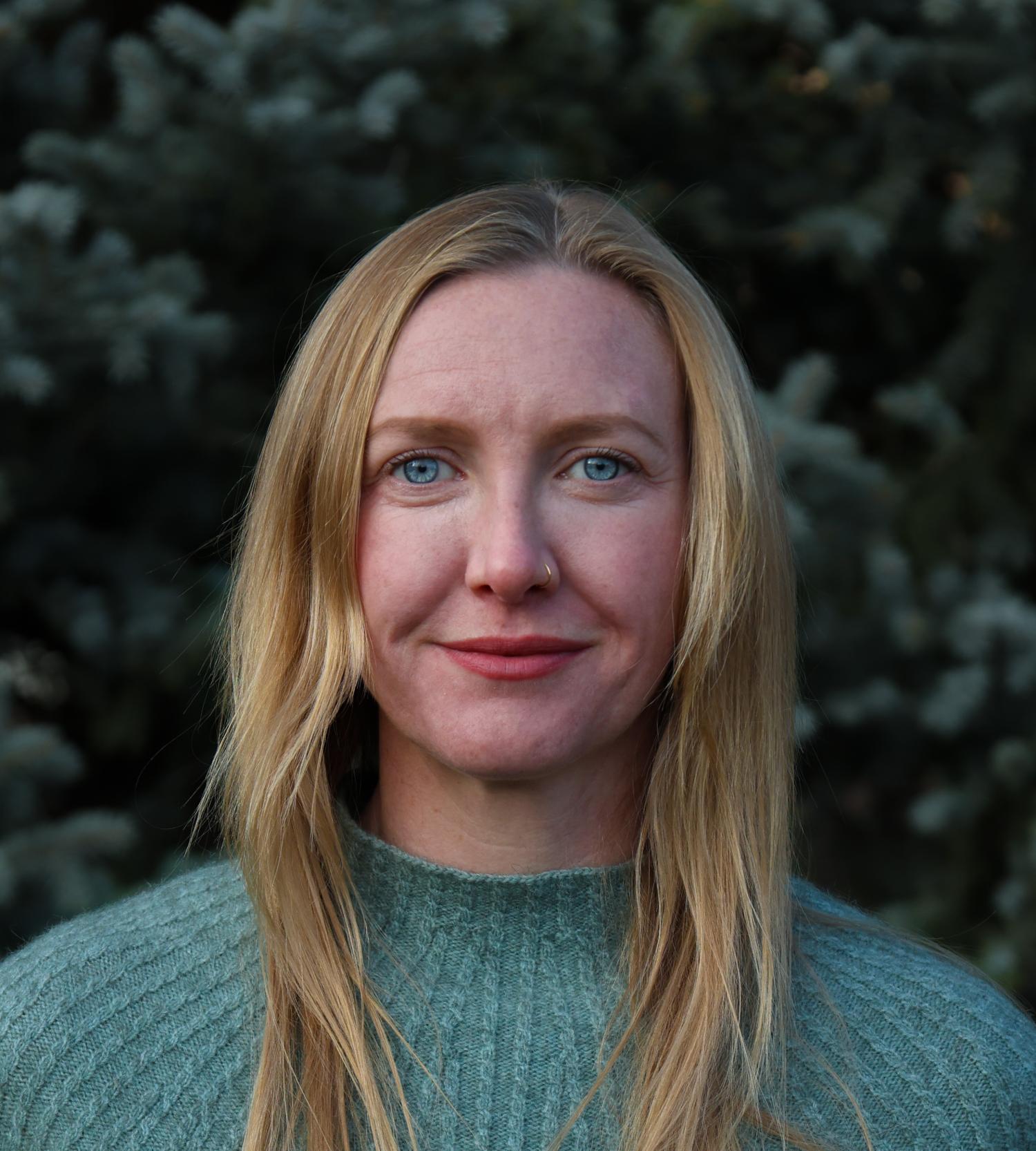 Woman with blonde hair and blue sweater stands in front of tree