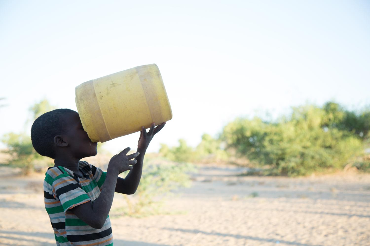 Boy in striped shirt drinking from a large container outside in the African landscape