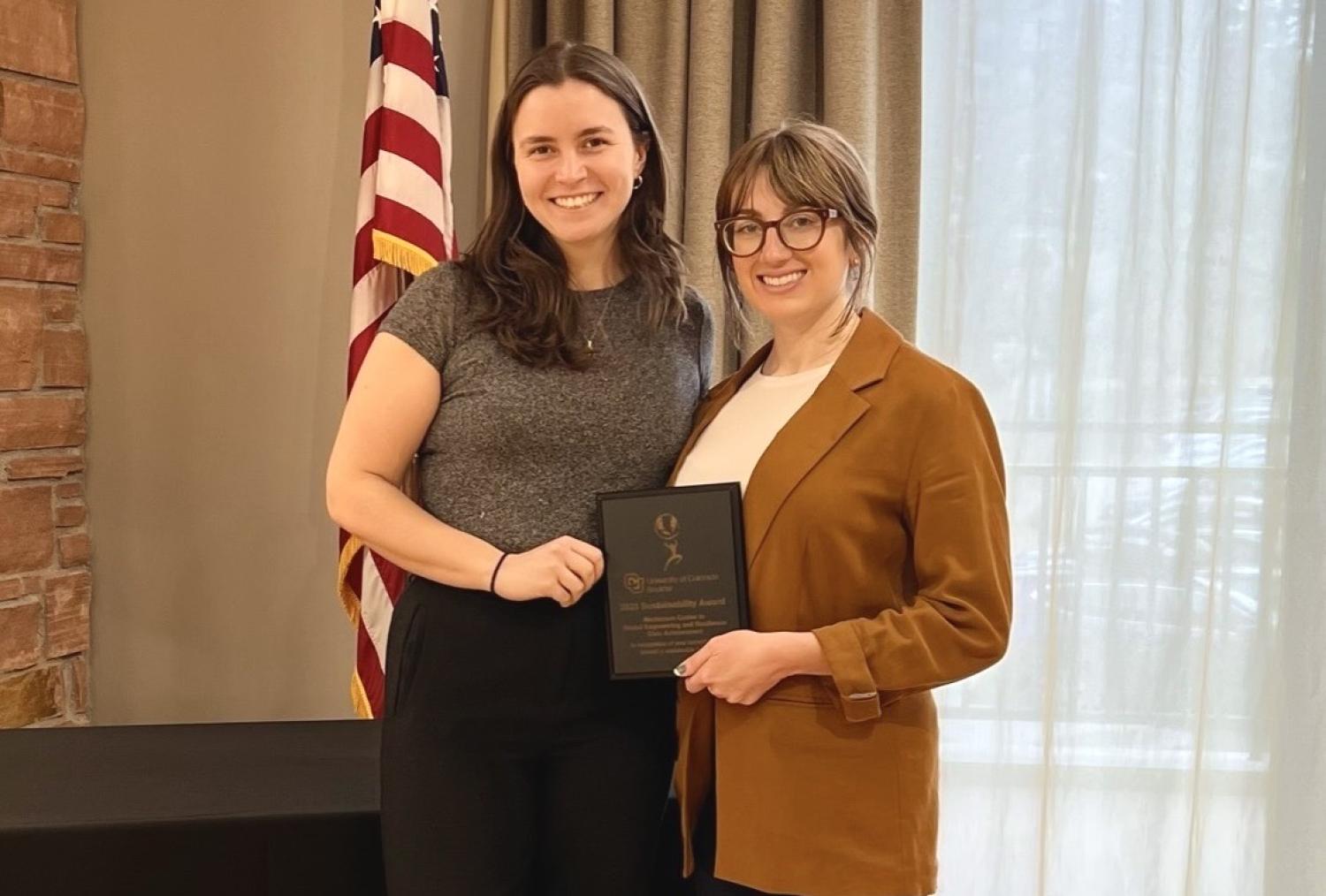Two women posing with an award plaque with a window and US flag in the background