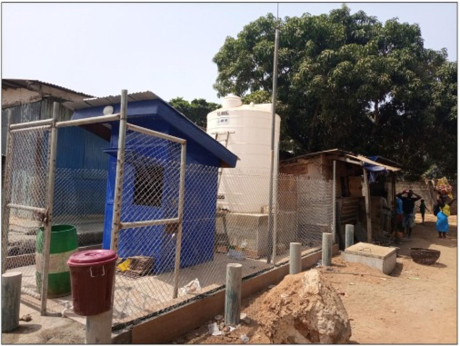 Small blue building with white storage tank behind a wire fence in a semi-informal settlement
