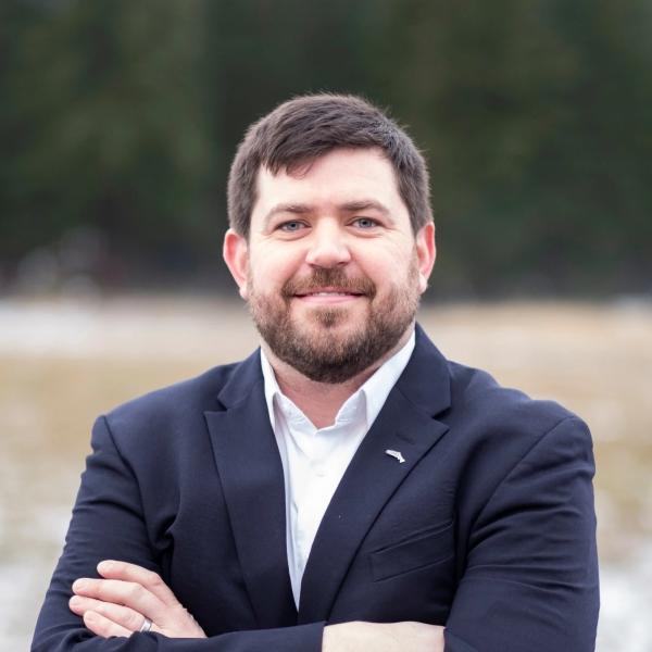 Man with a groomed beard and crossed arms smiling while wearing a black sports jacket 