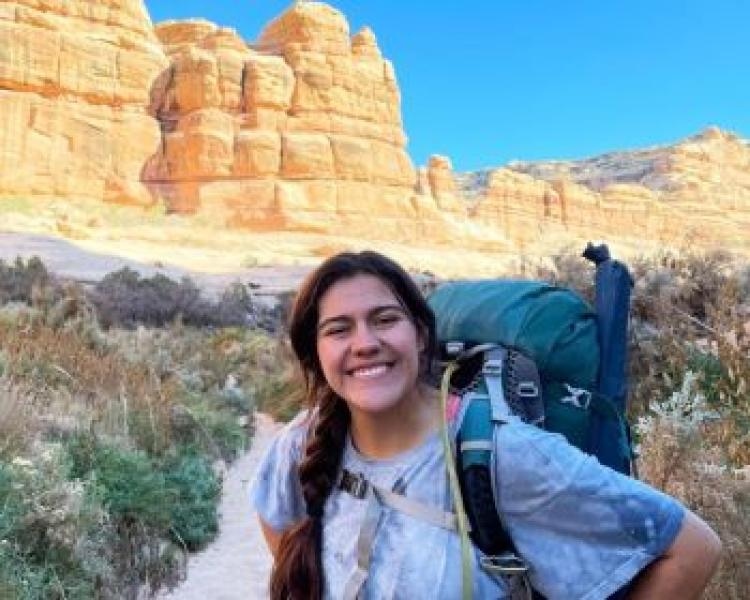 A person with dark hair in a braid wearing a backpack smiling at the camera standing outside in front of cliffs