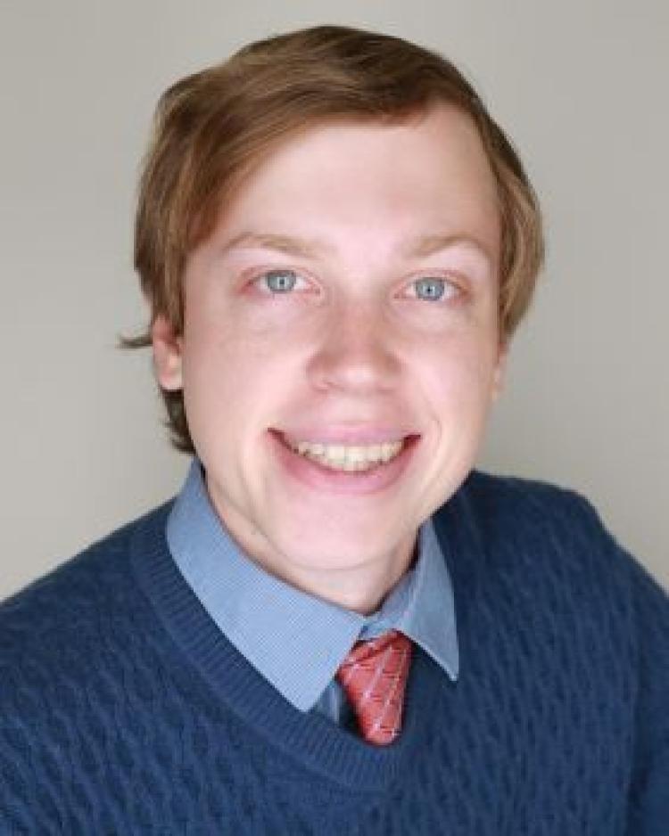 A person with short light brown hair wearing a dark blue sweater over a light blue collared shirt and pink tie smiling at the camera