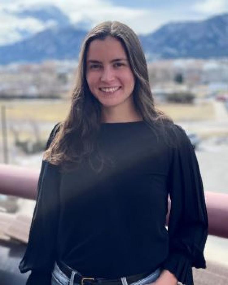 A person with long dark hair wearing a black top smiling at the camera standing outside on a balcony in front of mountains