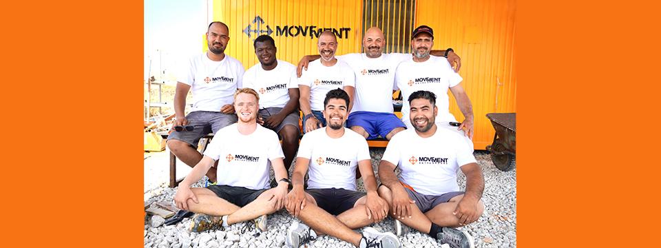 Eight men sitting cross legged on the ground and on a bench with white logo tshirts