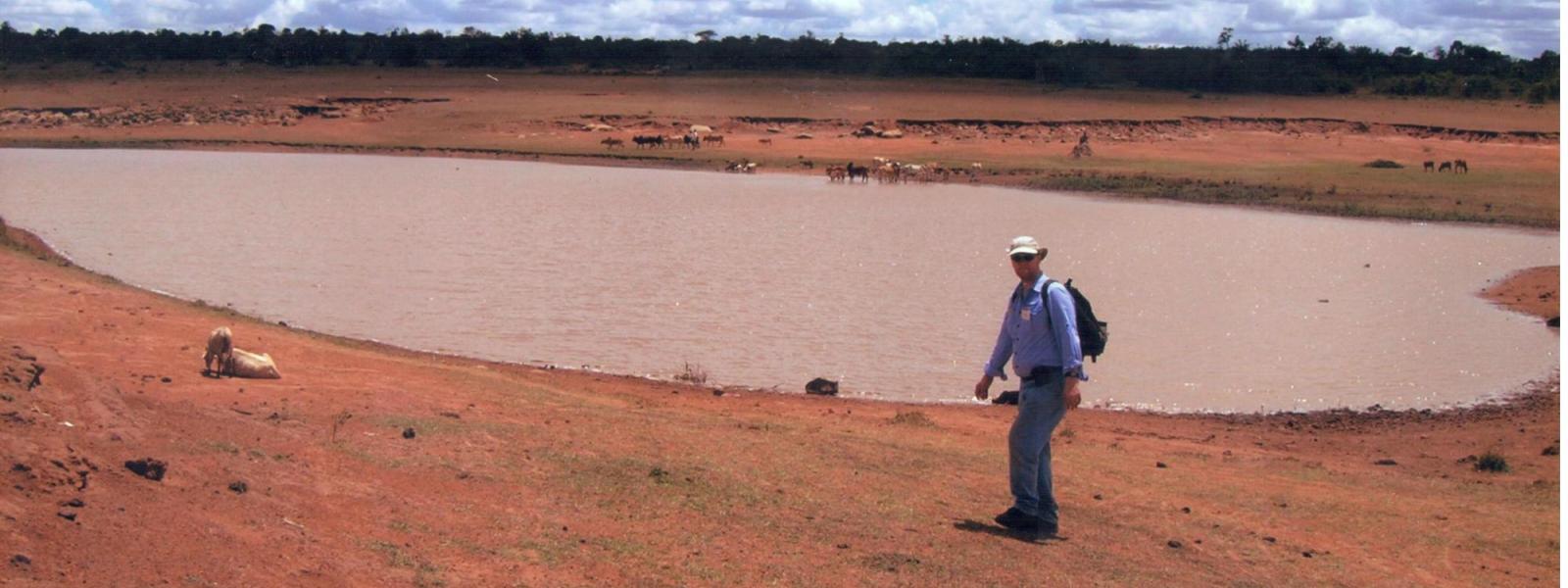 Man pauses in front of a large, dirty pond of water used as a source of drinking water with animals in background