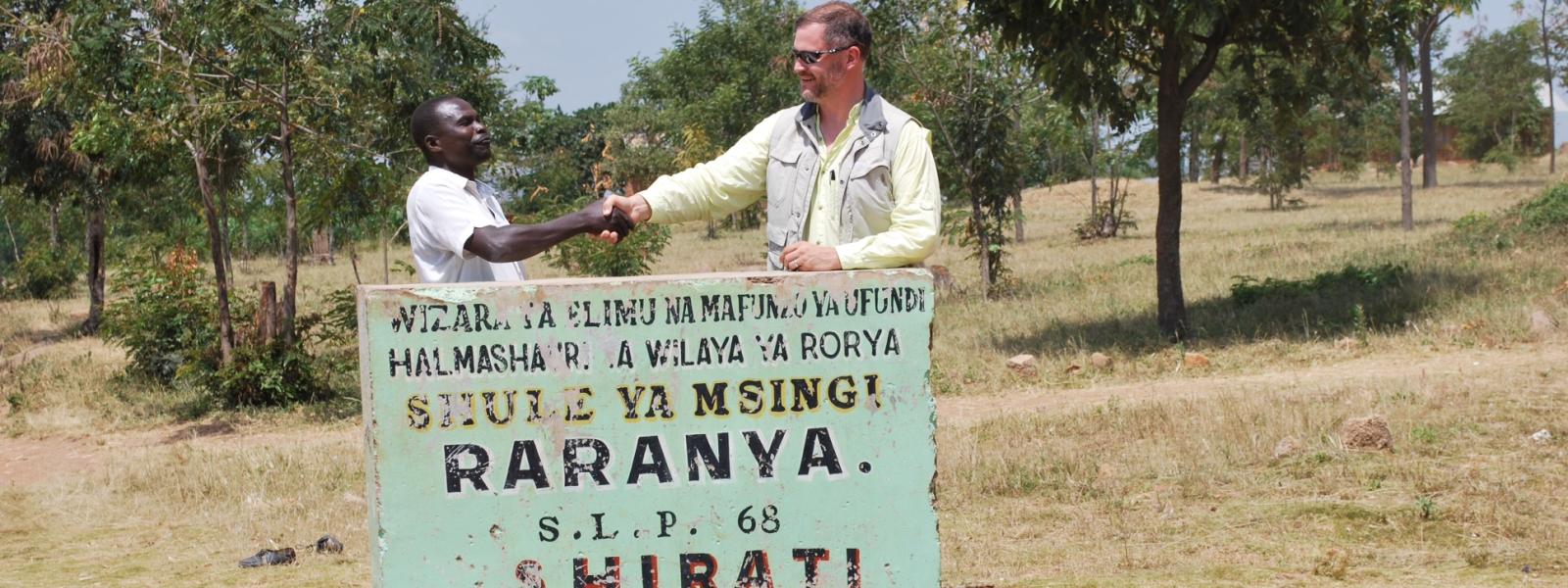 Man shaking hands with a Tanzania local in front of Raranya village's welcome sign