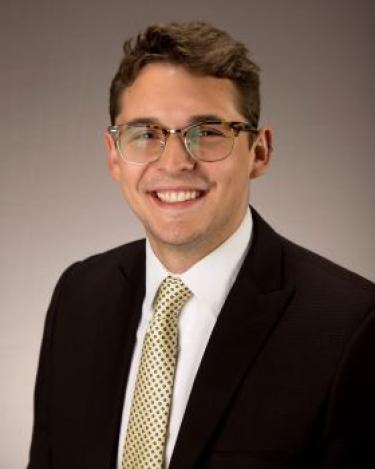 A professional headshot of a person with short dark hair wearing glasses and a suit smiling at the camera