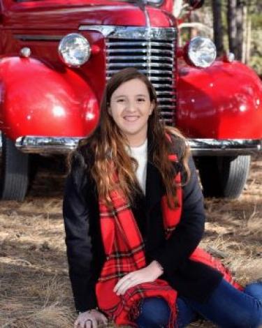 A person with long light brown hair smiling at the camera sitting on the ground outside in front of a red vintage car