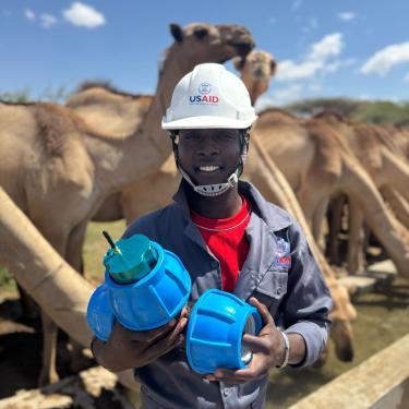 Team member holding a sensor in both hands outside in Kenya in front of camels.