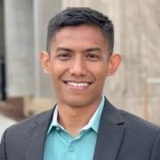 Student standing in a blue suit with blue shirt smiling at the camera 