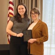 Two women posing with an award plaque with a window and US flag in the background