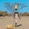 A person walking away carrying a yellow water container on their head while dragging another water container behind them