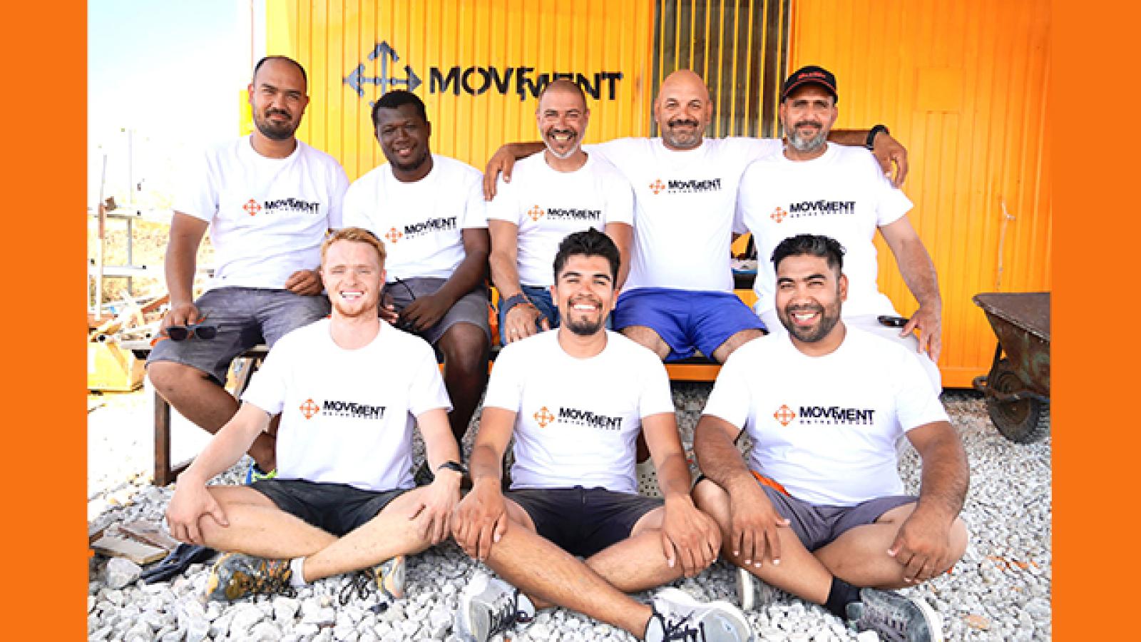 Eight men sitting cross legged on the ground and on a bench with white logo tshirts