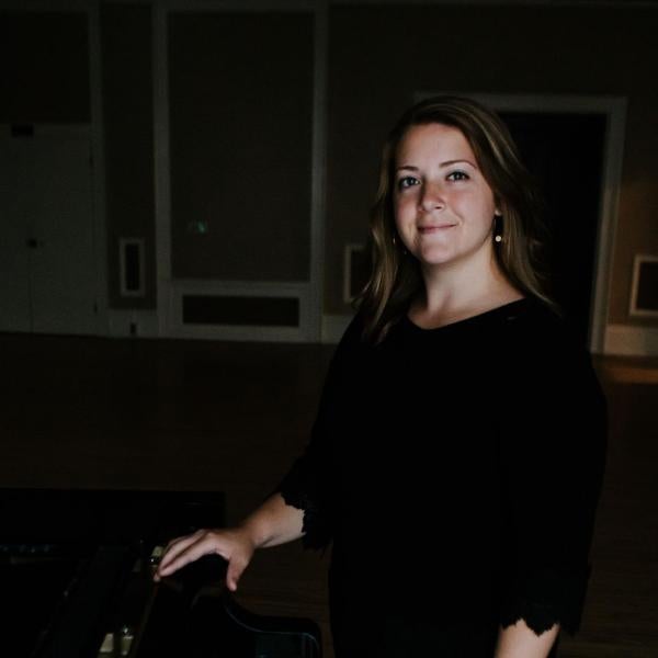 short blonde woman smiling and standing next to piano with hand on keys