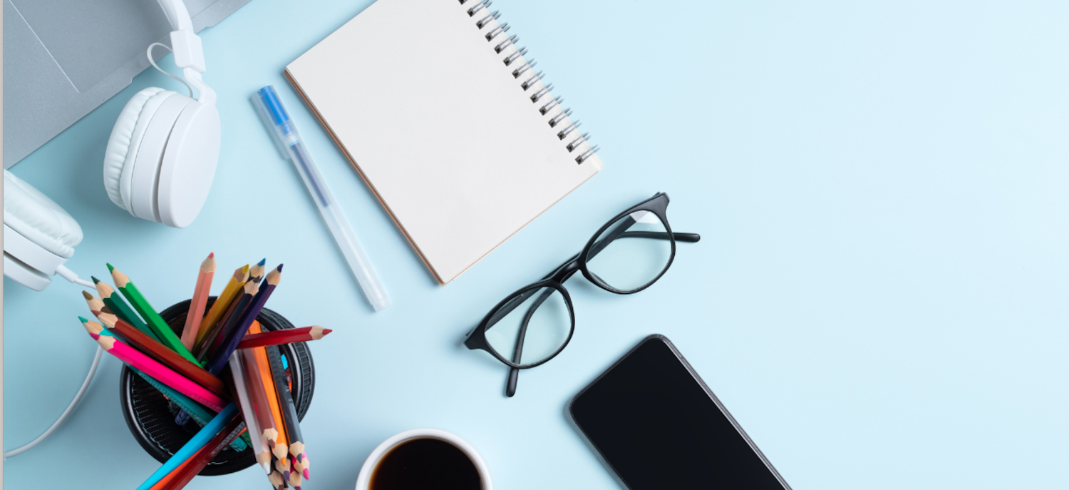 A surface with a computer, headphones, notebook, pen, colored pencils, phone, glasses, coffee, and post-it notes.