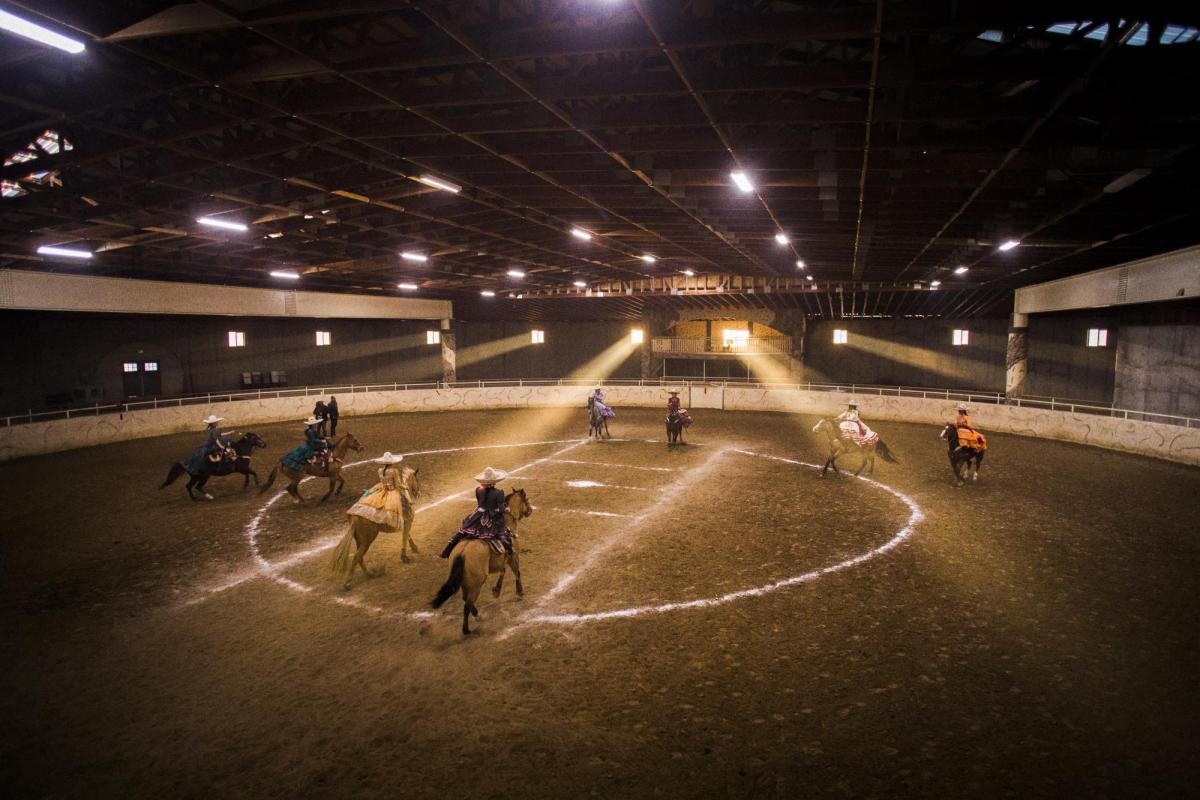 Escaramuza riders and horses practice in an indoor arena filled with beams of sunlight
