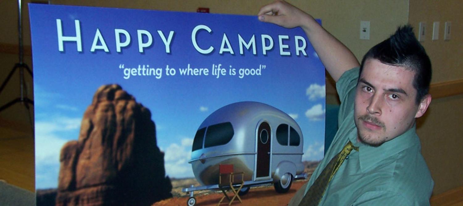 photo of Jesse Case holding a poster stating "Happy Camper, get to where life is good"