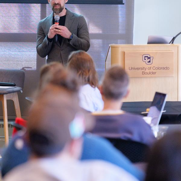 Caleb Elfenbein holding a microphone while presenting his work, audience is blurred