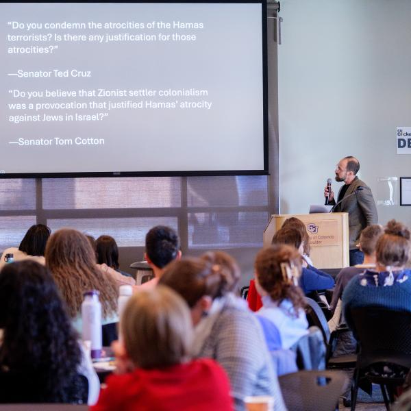 Caleb Elfenbein holding a microphone and presenting his work, with powerpoint behind him; audience is shown