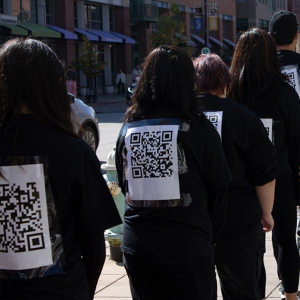 Group of people from political art performance is seen wearing a black shirt; back shown is a QR Code. The QR code, if scanned, corresponds to landmark civil rights case.