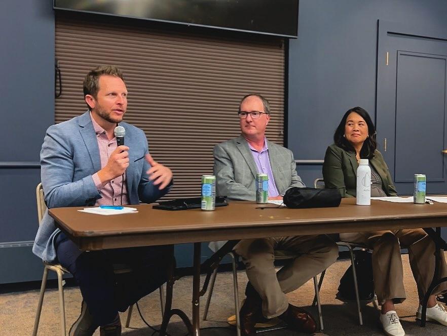 A man in a blue blazer holding a microphone sitting down, speaking to an unseen audience, with a man and a woman on his left looking at him. They are sitting at a table