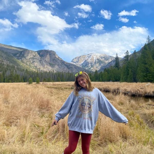 Cora Becker in a sweatshirt with arms outstretched in a field with mountains in the background.