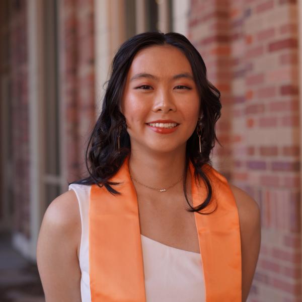 Judy Chen in a white dress with an orange graduation scarf in front of a blurred sandstone brick building.