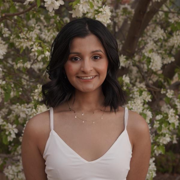 Tanvi Patil in a white sleeveless dress in front of a flowering tree.