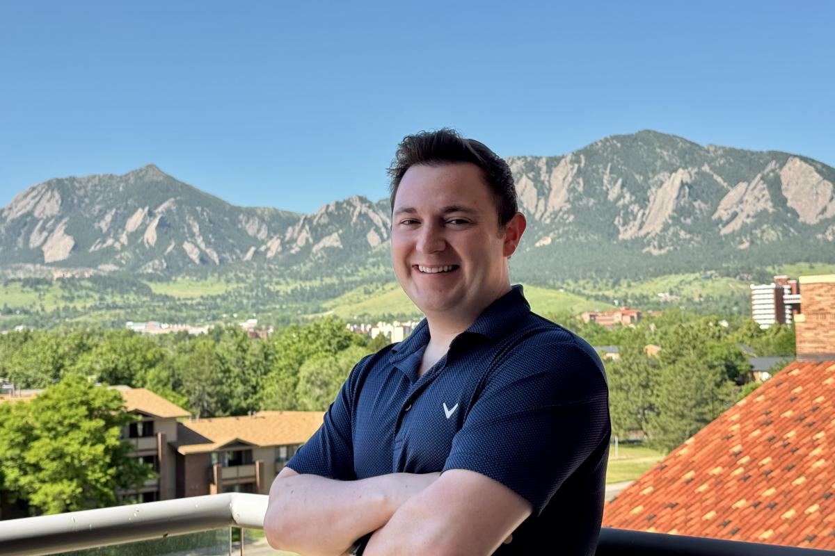 Nolan Petrich in a short-sleeved polo with his arms crossed and part of a roof and the Flatirons in the background