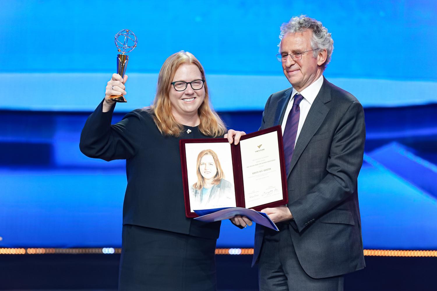 Kristi Anseth holds up her award at the 2024 VinFuture Prize Award Ceremony with a folder open to her award and a sketch of her held by Sir Richard Henry Friend.