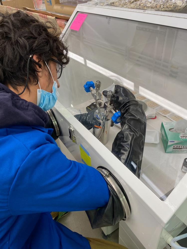 Barrientos wearing a blue lab coat, safety glasses, and a face mask works with scientific equipment inside a glove box in a laboratory setting. His arms are inserted into black rubber gloves attached to the sealed chamber, which contains metal instruments and lab tools.