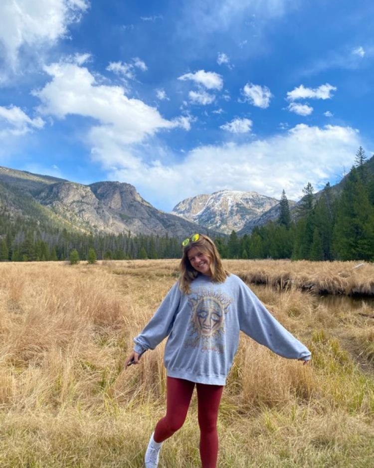 Cora Becker in a sweatshirt with arms outstretched in a field with mountains in the background.