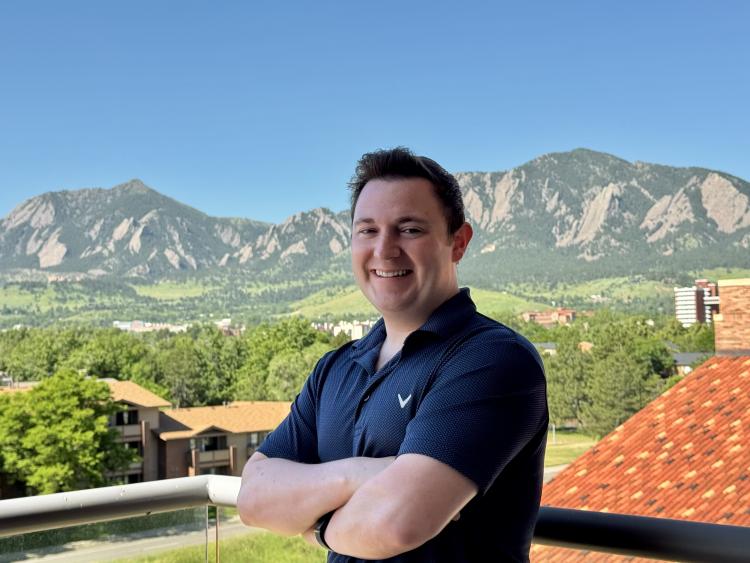 Nolan Petrich in a short-sleeved polo with his arms crossed and part of a roof and the Flatirons in the background