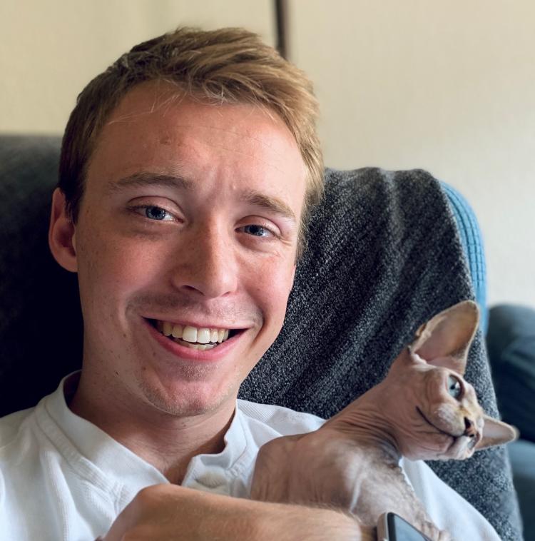 Bruce Kirkpatrick smiling while sitting on a couch, holding a hairless Sphynx cat that looks off to the side. A textured gray blanket is draped over the back of the couch.