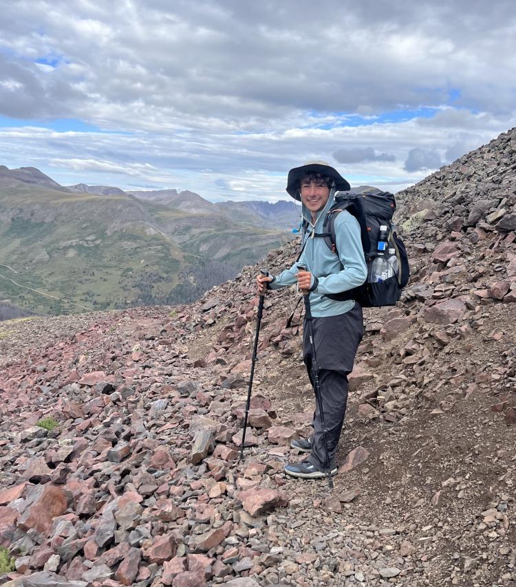 Ethan Coleman wearing a wide-brimmed hat, light blue jacket and large backpack stands on a rocky mountain trail, using trekking poles. Behind him are expansive green valleys and distant mountain peaks under a partly cloudy sky.